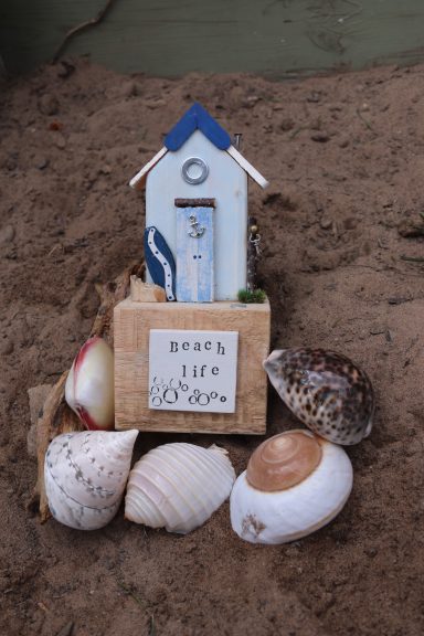 A decorative beach-themed birdhouse surrounded by various seashells on sand.