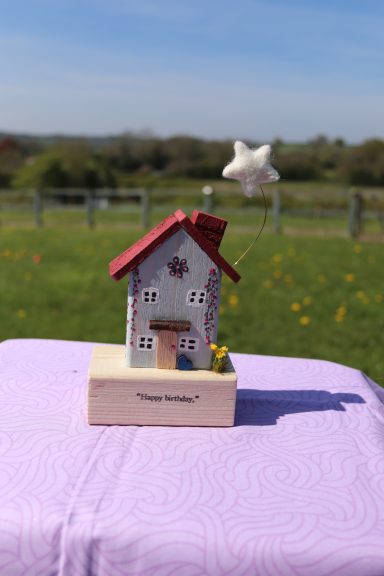 A decorative miniature house on a wooden base with a flower, set in a grassy field.